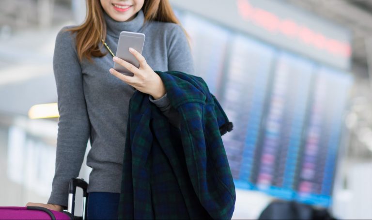 Charming beautiful business woman smile in casual style using smartphone and hold luggage bag for travel with blur the airport terminal.